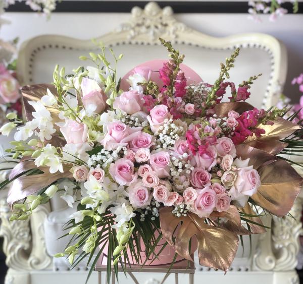 Rose gold gilded tropical leaves surround this beautiful arrangement of white Orchids, pink roses, spray roses, and Snapdragons. Highlighted with mixed tropical greens and made in a large hat box.